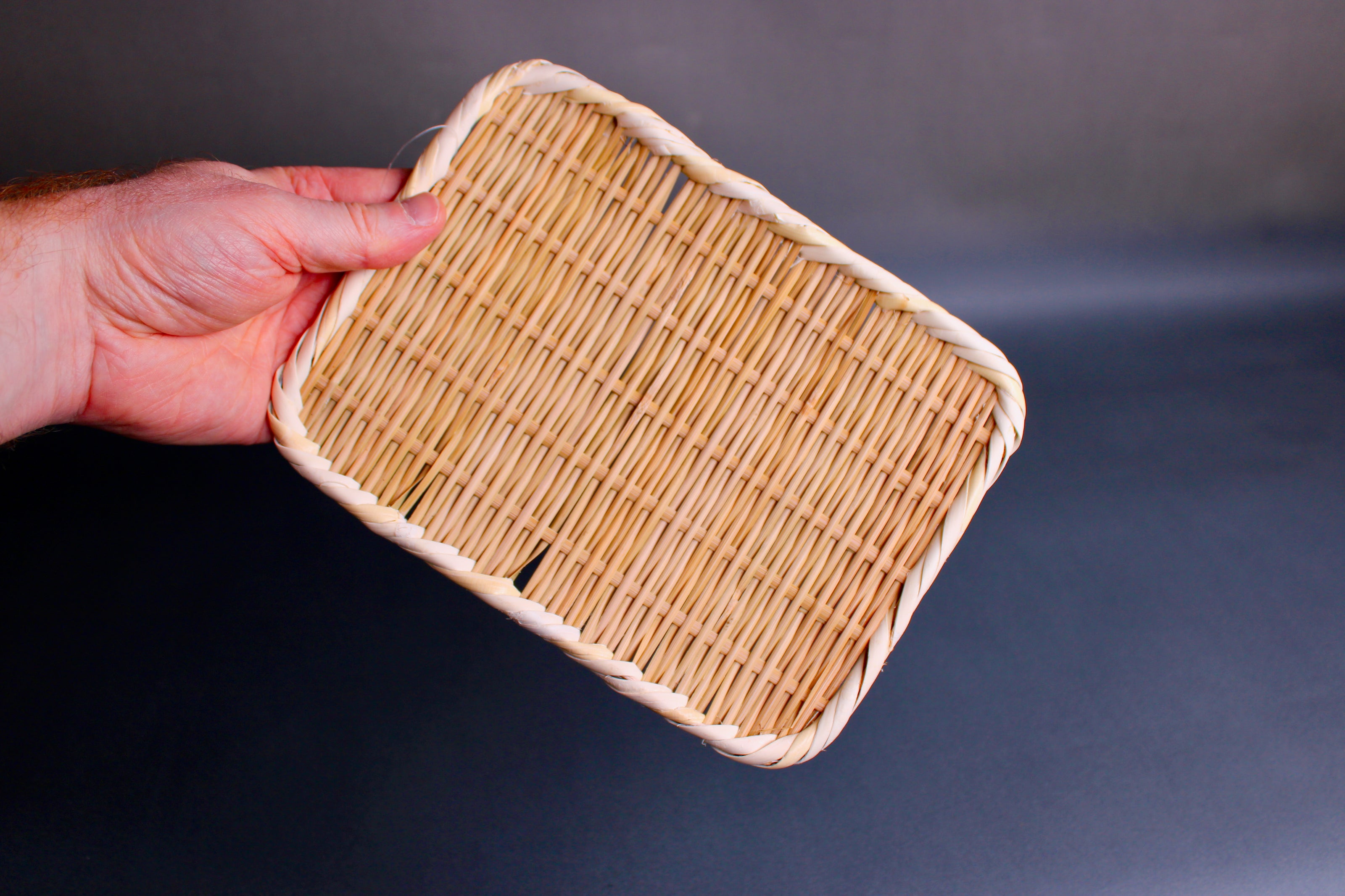 hand holding woven bamboo strainer rectangular tan brownish in color in grey blue backdrop