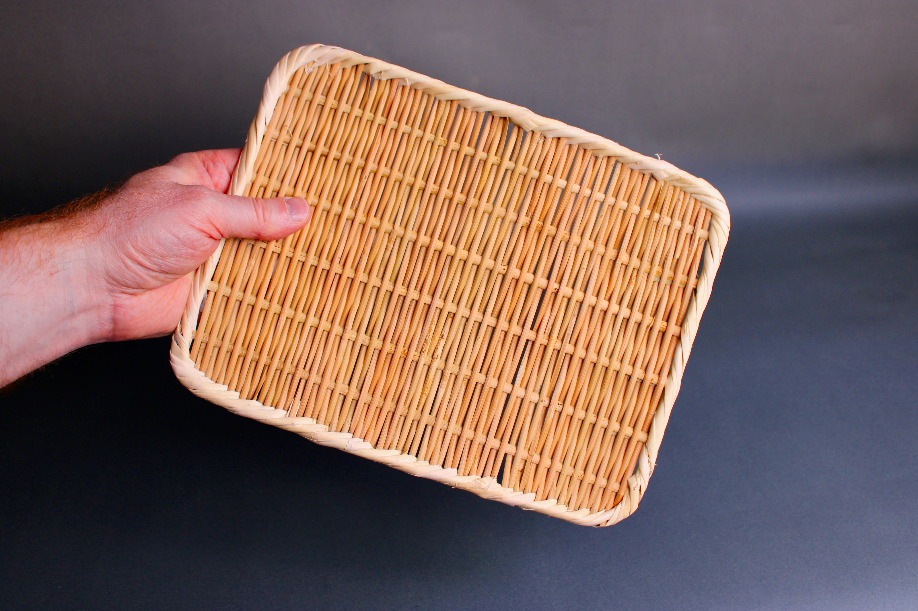 hand holding woven bamboo strainer rectangular tan brownish in color with grey blue backdrop