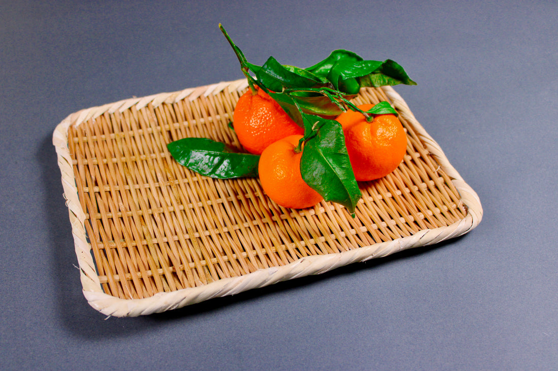 woven bamboo strainer rectangular tan brownish in color bright orange satsuma atop with green leaves in grey blue backdrop