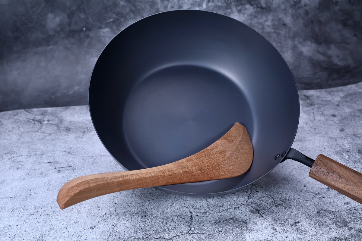 Blue carbon steel wok with wooden handle and wooden spatula resting inside, angled view on stone surface with grey background