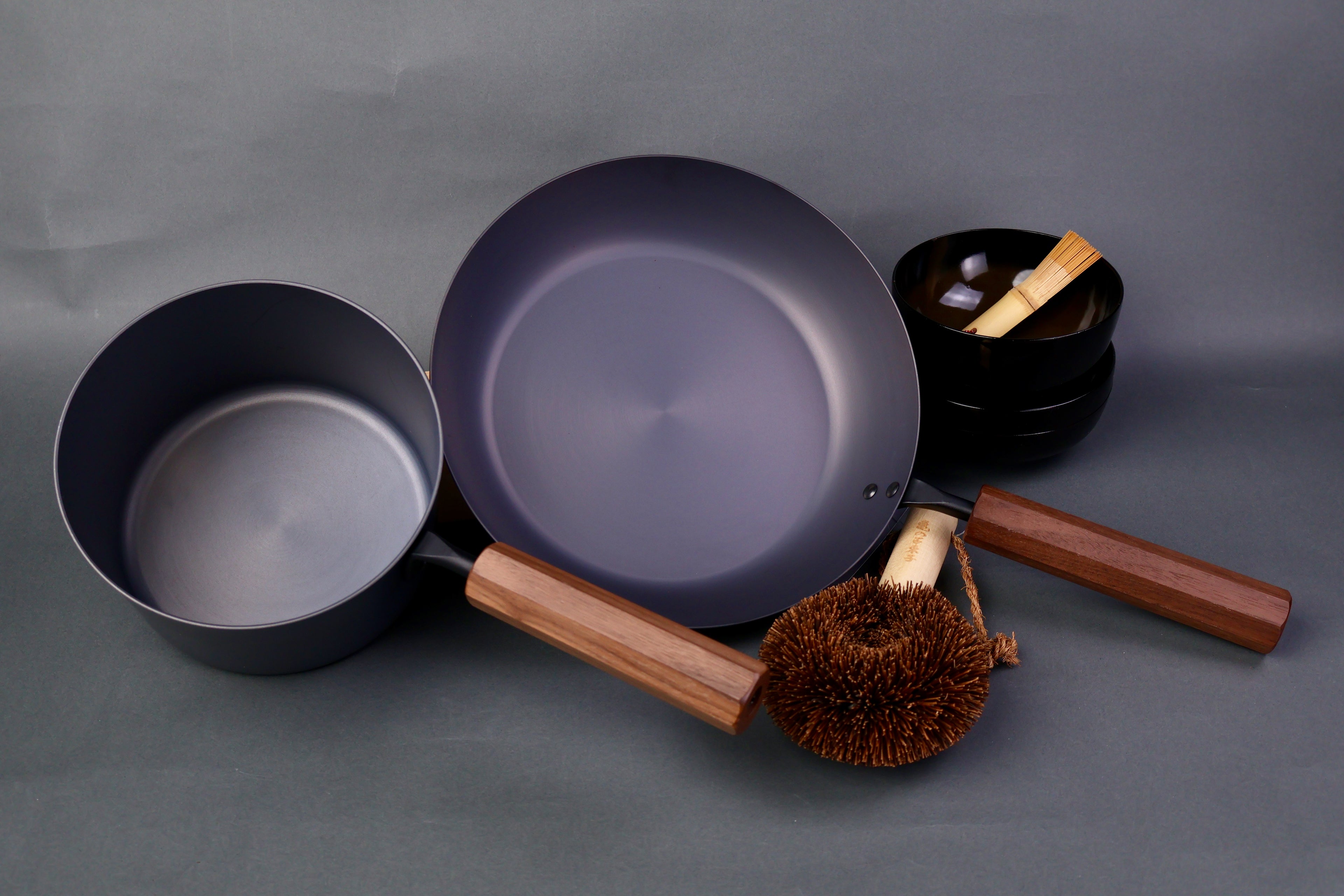 wide low bluish grey black carbon steel saute pan next to tall narrow dark grey iron saucepan both with octagonal wood handle in grey background