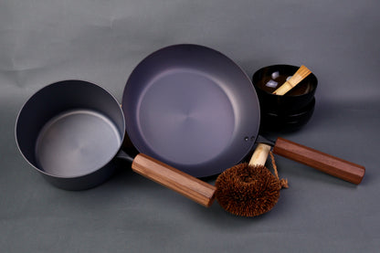 wide low bluish grey black carbon steel saute pan next to tall narrow dark grey iron saucepan both with octagonal wood handle in grey background
