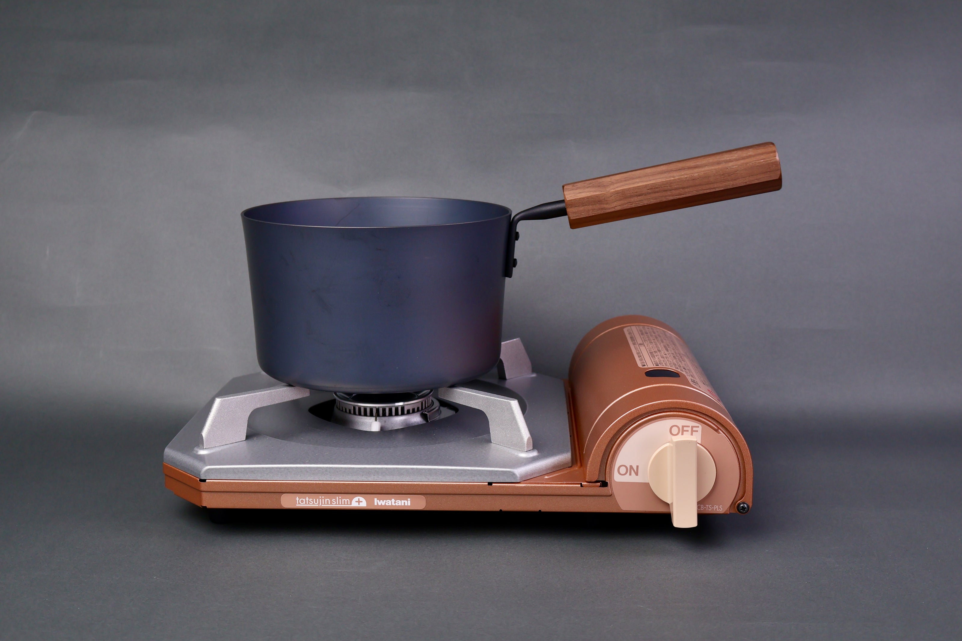 dark grey carbon steel tall narrow iron saucepan with wood handle sits atop portable butane stove  in light grey background