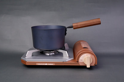 dark grey carbon steel tall narrow iron saucepan with wood handle sits atop portable butane stove  in light grey background