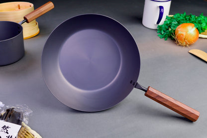frontal shot of grayish black nitrided iron saute pan with brown wooden octagonal handle surrounded by Japanese kitchenware with light grey background
