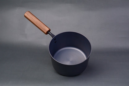 dark grey carbon steel tall narrow iron saucepan with octagonal deep brown wood handle in light grey background