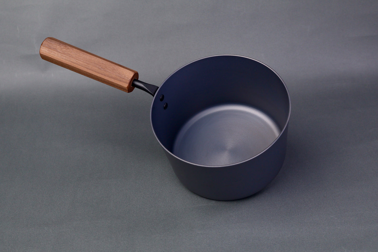 dark grey carbon steel tall narrow iron saucepan with octagonal deep brown wood handle in light grey background