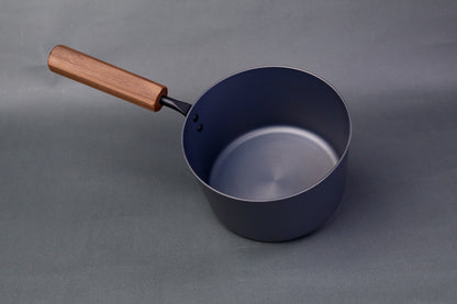 dark grey carbon steel tall narrow iron saucepan with octagonal deep brown wood handle in light grey background