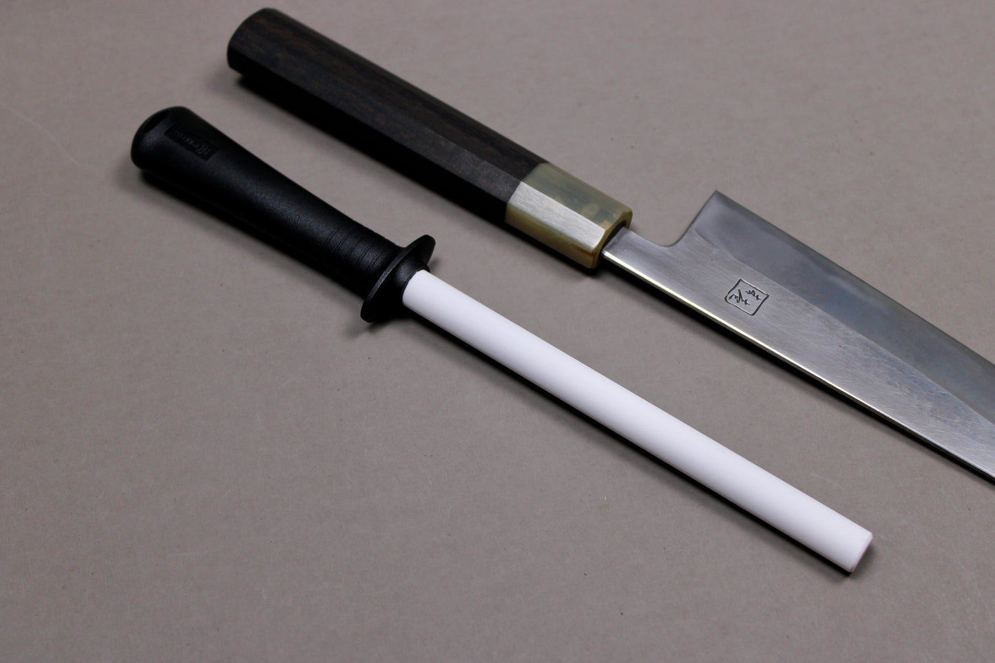 plastic black handled white ceramic honing rod angled next to wood handled steel knife with kanji engraving on grey background