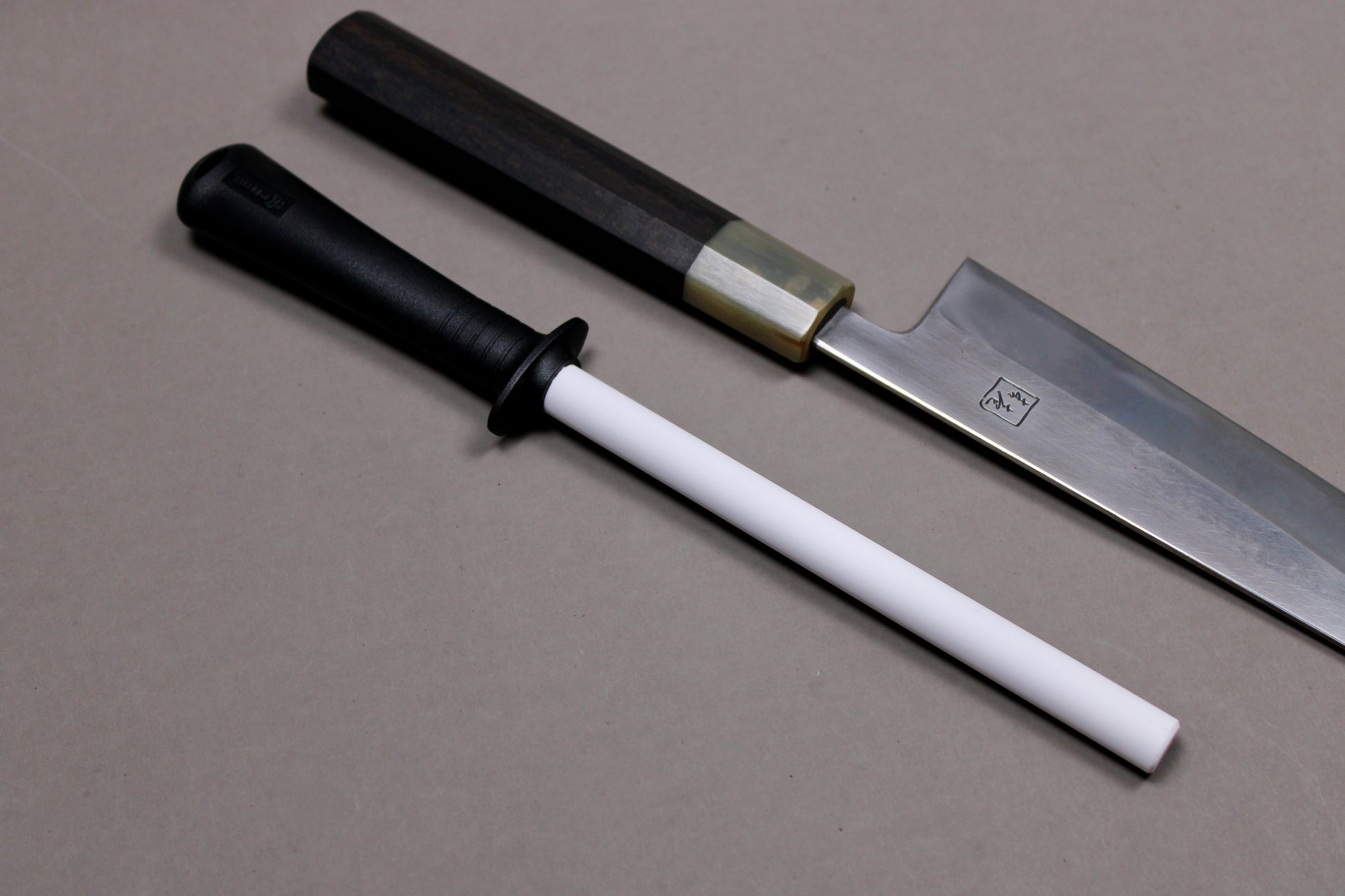 plastic black handled white ceramic honing rod angled next to wood handled steel knife with kanji engraving on grey background