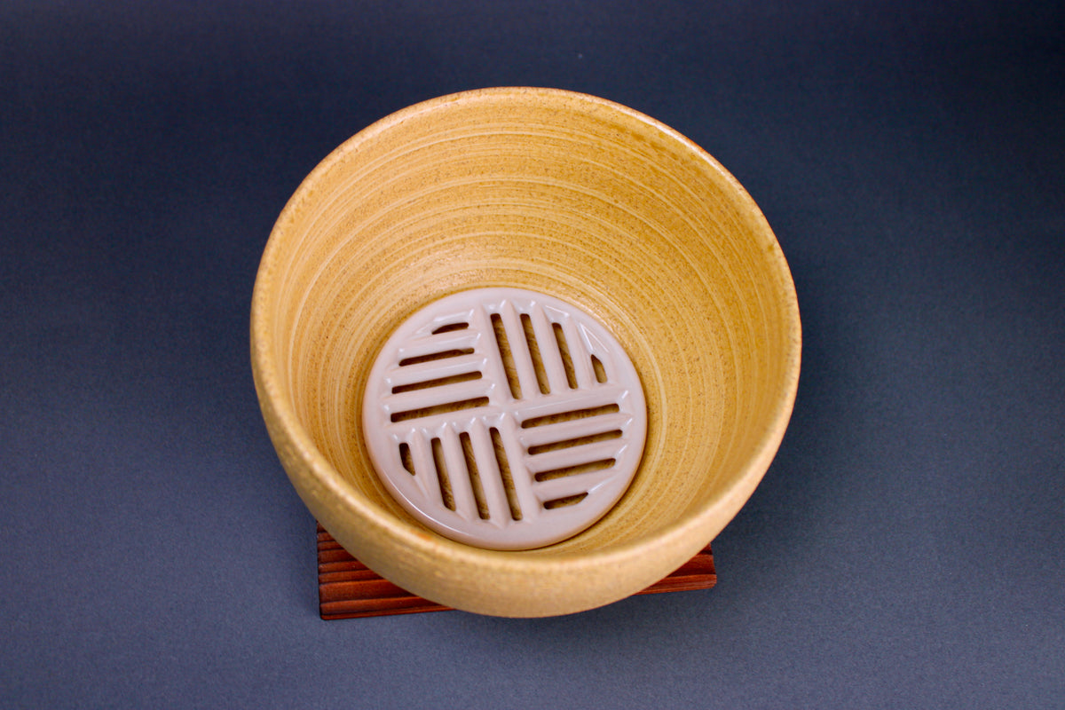 short round yellowish ceramic bowl with white grate inside sitting atop tan wood square trivet in grey background