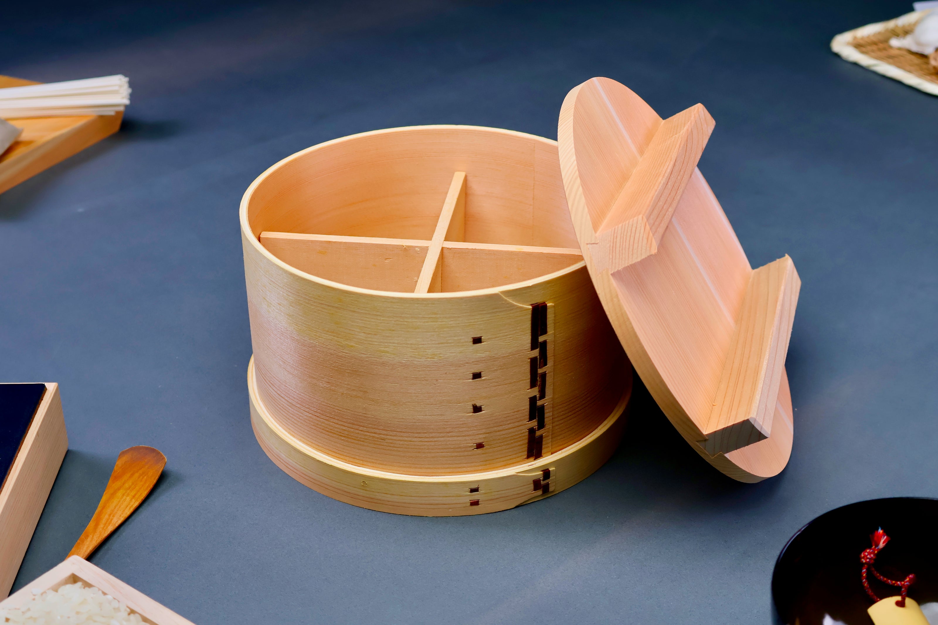 round natural wooden food steamer two handled round lid leaning against body surrounding japanese kitchenware  in grey background  