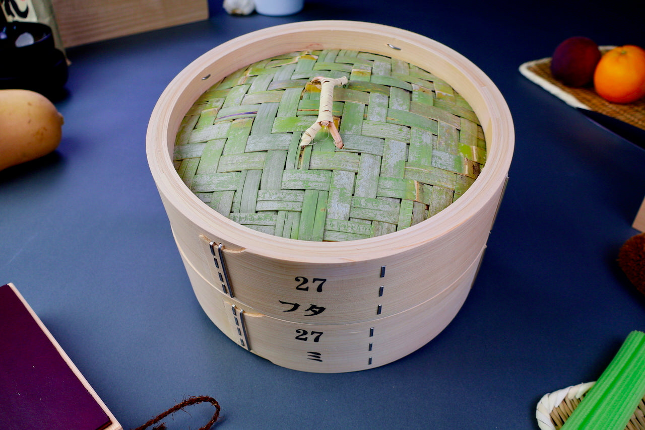 thick wood round bamboo steamer set woven top with loop natural wood tone with printed hiragana surrounded by japanese kitchenware blue gray background