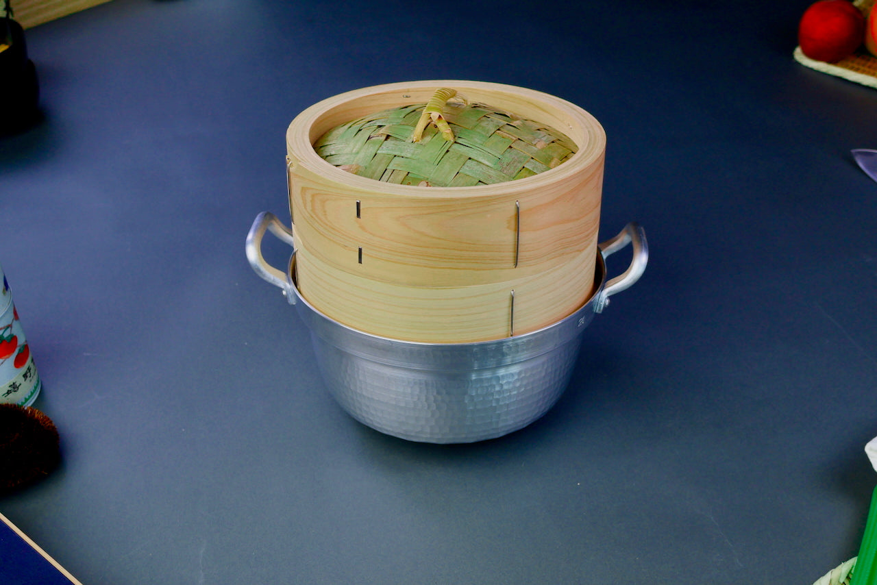 top down shot of round hinoki wood steamer with lid and base neatly fitted in silver hued two handle saucepan grey background