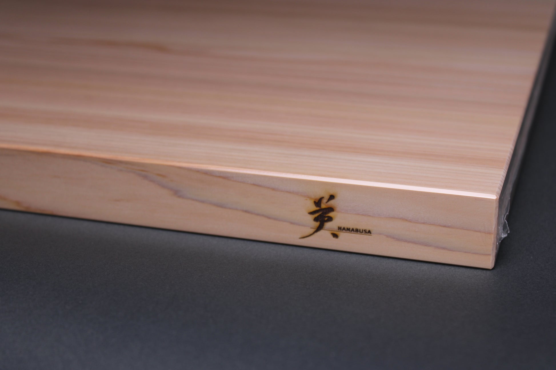 detailed close up shot of hanabusa logo written in English next to kanji logo of the same brand the detailed grain pattern of the wood is shown on the front face of the board and details of the chamfered edges are shown along with the creamy wood texture of the board in front of dar grey background 