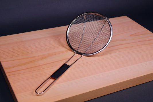 Metal round strainer with long thin handle on a hinoki cutting board with a dark background