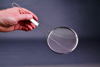 Metal coarse skimmer with long thin handle held by a hand against a dark background