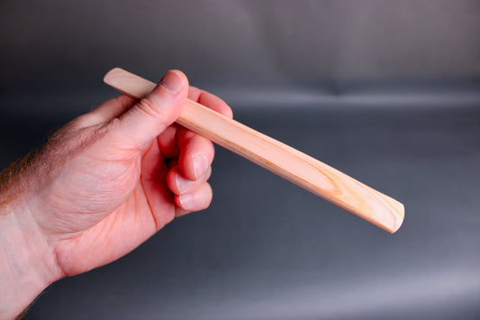 hand holding long thin flat natural wood tone hinoki spreader in dark grey backdrop 
