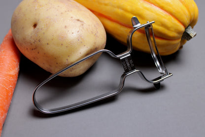 shimomura stainless curved vegetable peeler leaning against yellow potatoes and carrots with gray background