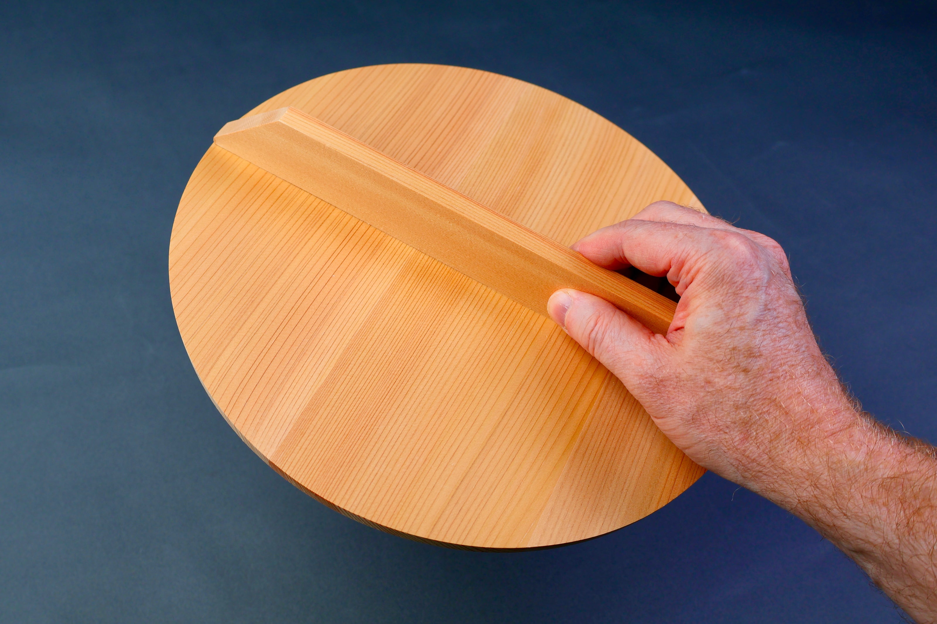 hand holding wooden circular lid with long shallow handle in front of bluish grey background
