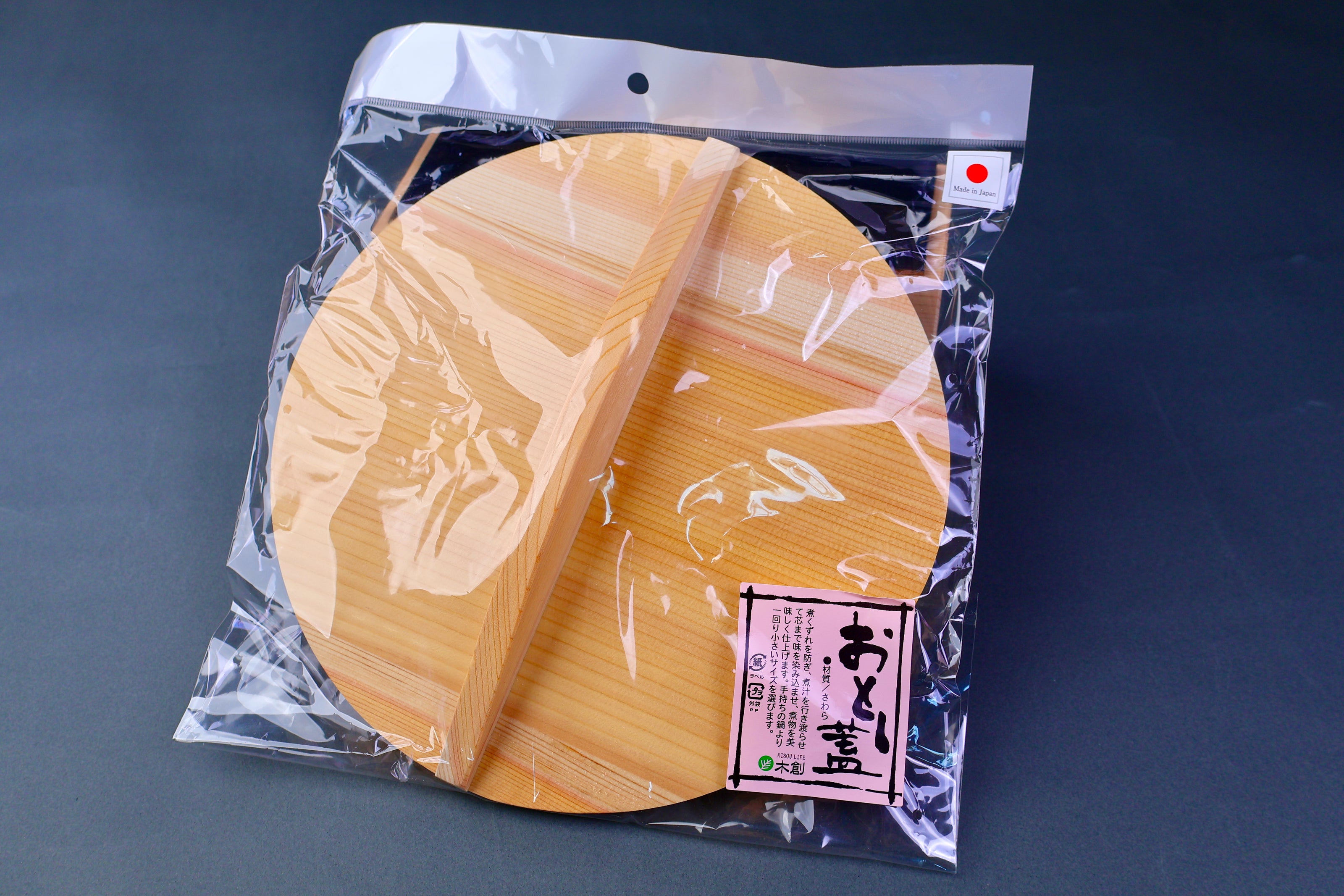 clear plastic package holding round wooden drop lid with long shallow handle in bluish grey background