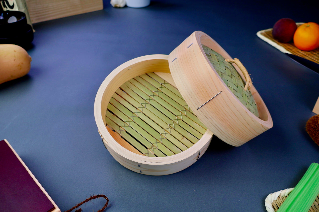 thick wood round bamboo steamer set woven top with loop natural wood tone with printed hiragana surrounded by japanese kitchenware blue gray background