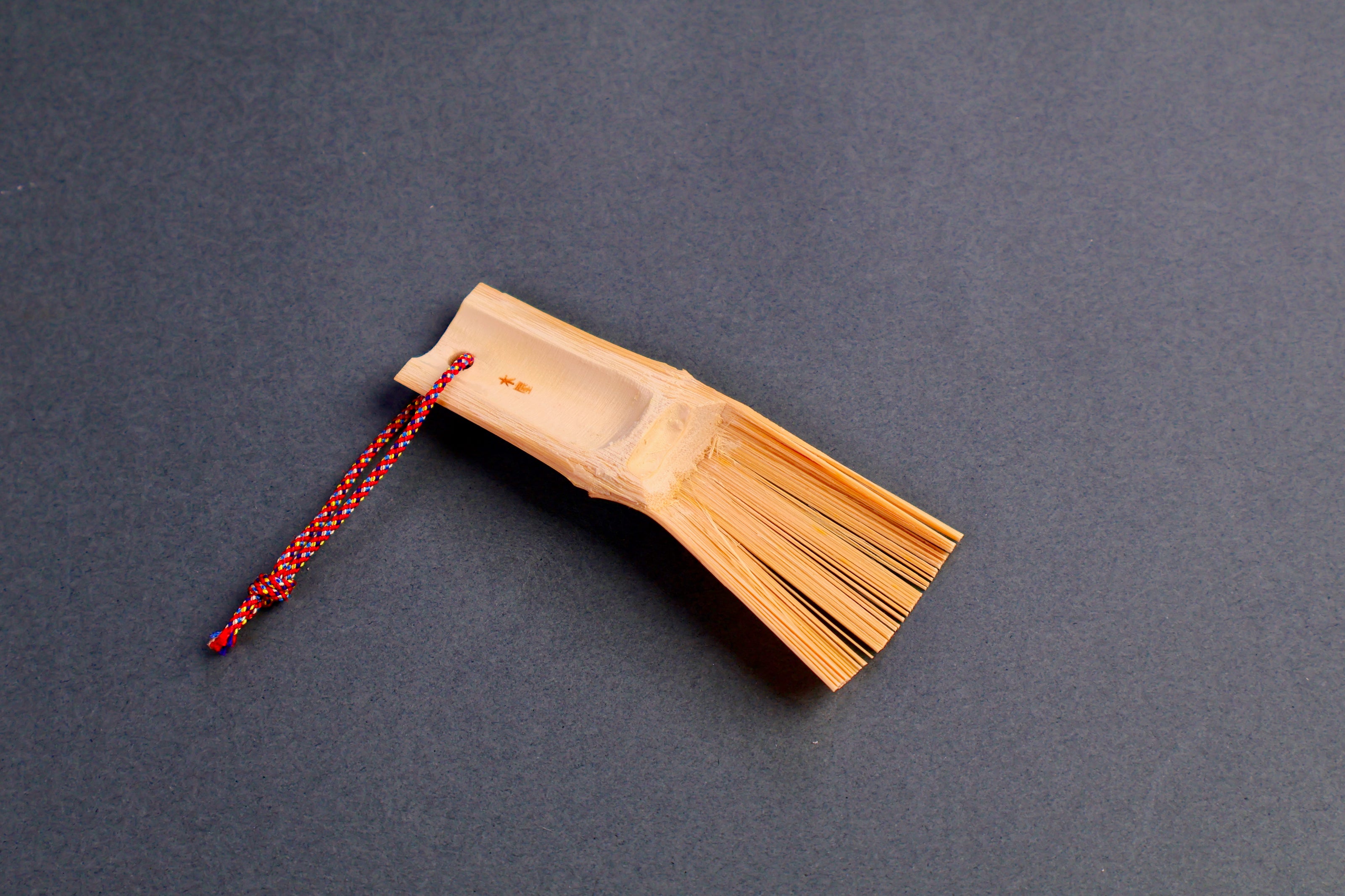 short rectangular bamboo brush with thin bristles red silk loop with tiny kanji print on dark grey backdrop
