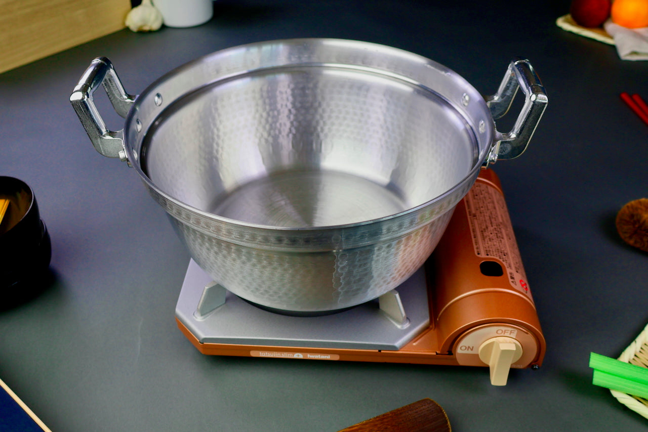 two handled stepped aluminum silvery pot with shiny hammered surface atop portable sliver tan butane stove surrounding kitchenware on a dark grey backdrop 