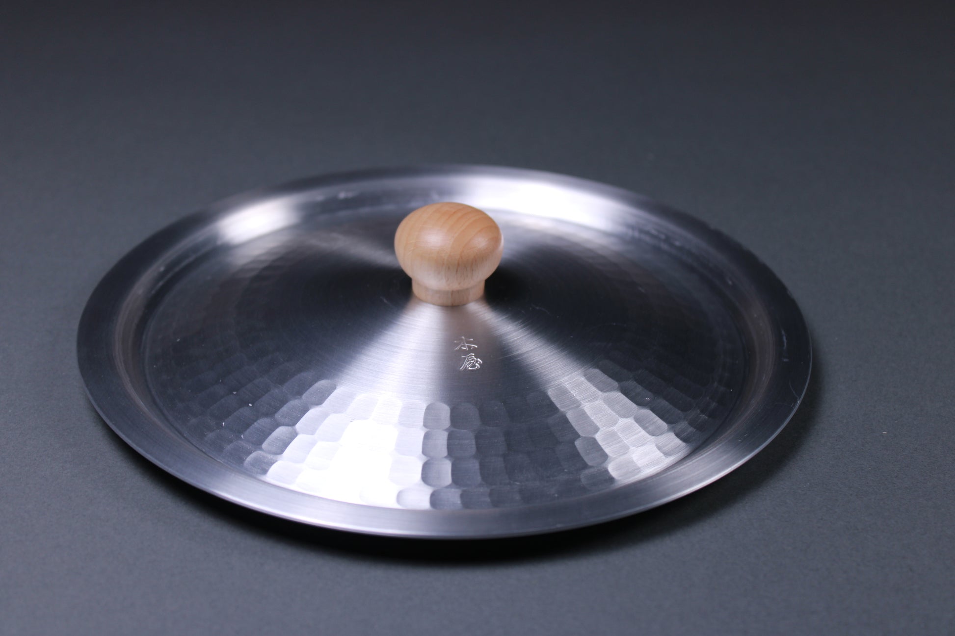 round metallic lid with hammered interior portion with wood knob in center sits on gray surface