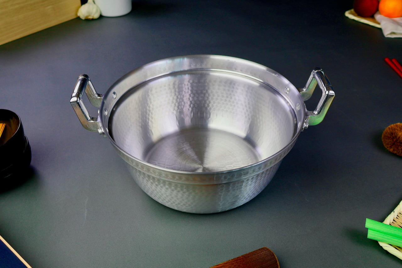 two handled stepped aluminum silvery pot with shiny hammered surface surrounded by japanese kitchenware on a dark grey backdrop 
