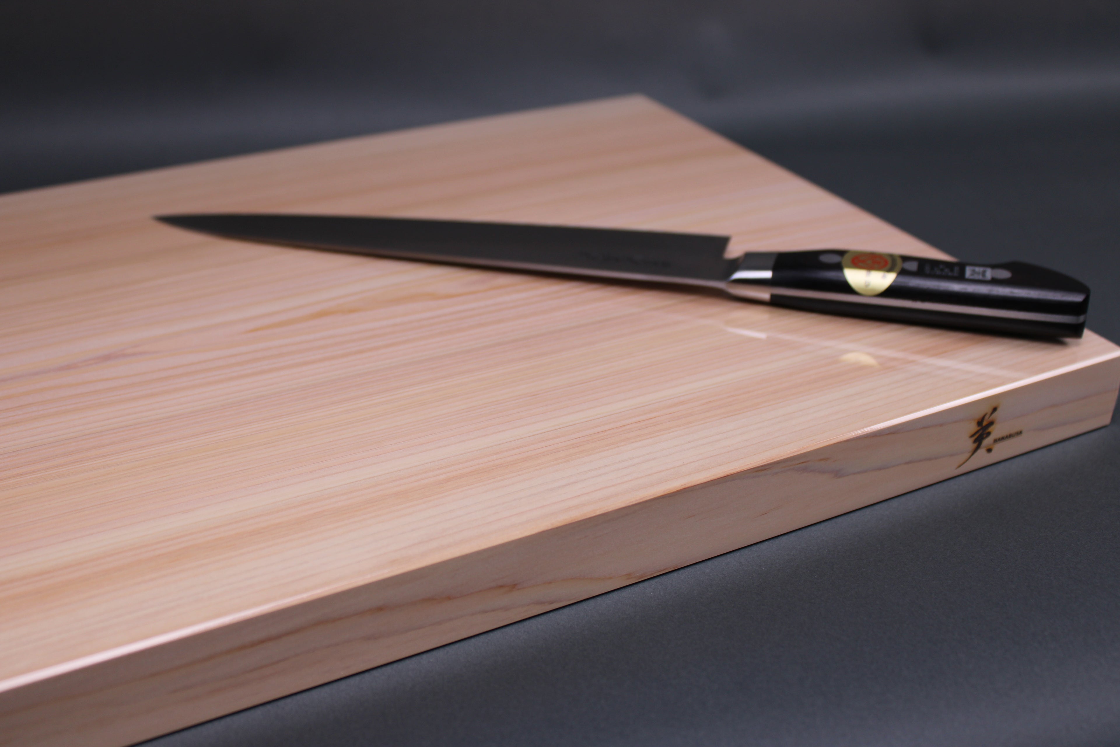 rectangular shaped light medium deep wood tan hinoki cutting board with deep natural grain lines black handled knife atop board in front of dark grey backdrop