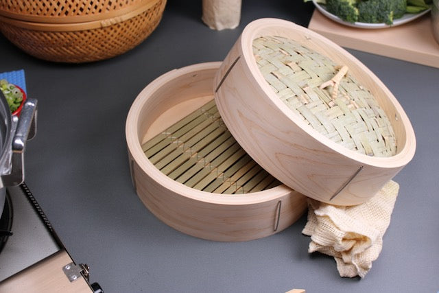  top down view of chinese bamboo and hinoki steamer laid open showing bamboo base and slats wrapped with stainless steel wire extra thick and durable japanese kitchenware surrounded by portable gas stove bamboo woven basket hinoki wood cutting board ceramic plate of sliced broccoli green cabbage next to white cotton steaming cloth