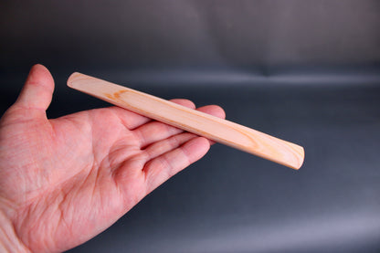 hand holding long thin flat natural wood tone hinoki spreader in dark grey backdrop 