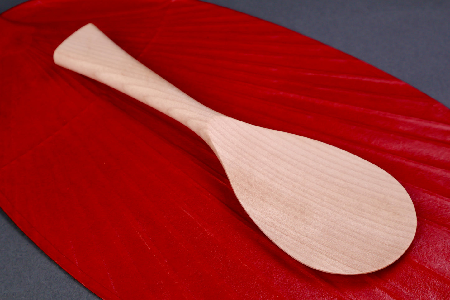 hand carved wooden rice paddle light golden brown yellowish tone of paddle atop deep red background surface