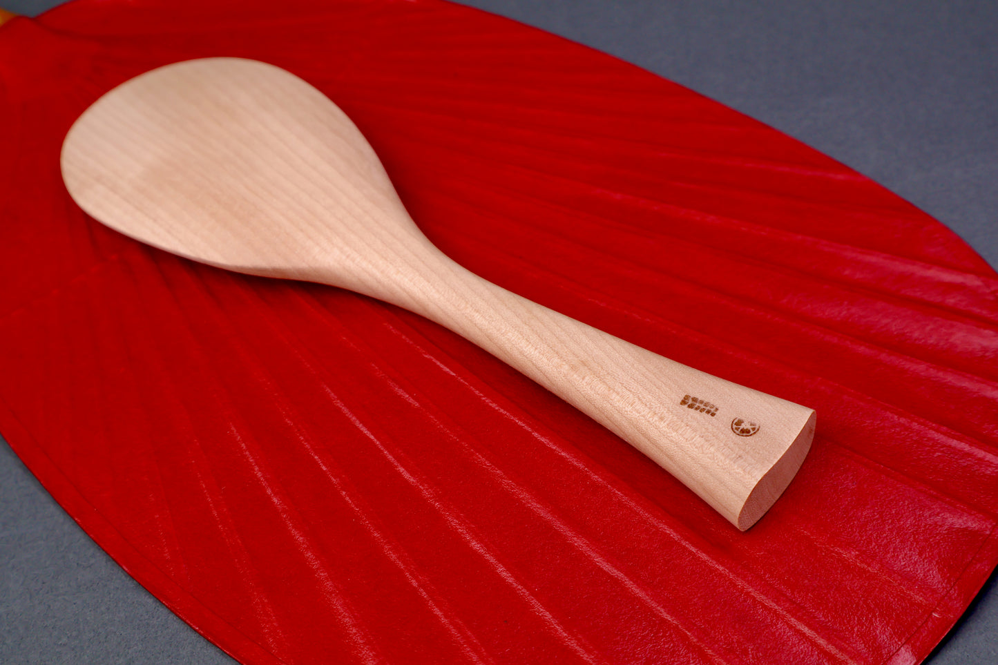 hand carved wooden rice paddle light golden brown yellowish tone of paddle atop deep red background surface