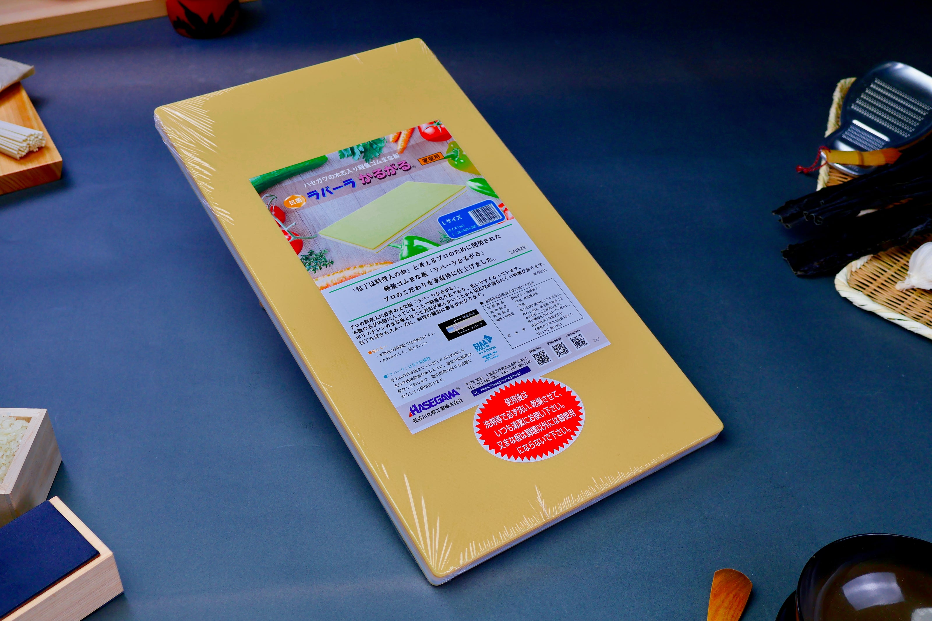 long yellow rectangular shaped rubber cutting board with product paper in hiragana showing bright green red blue images surrounded by kitchenware on grey background