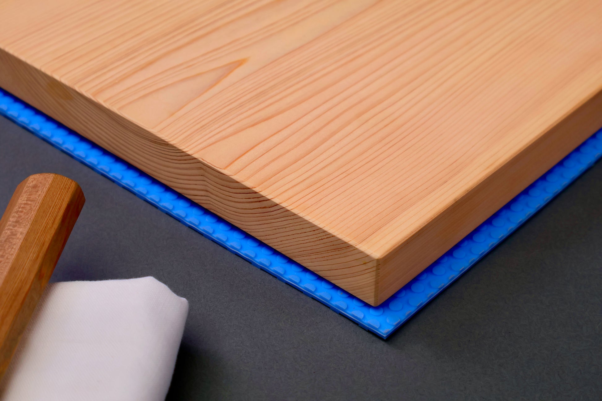 detail of natural wood tone hinoki cutting board corner sitting atop bright blue non slip rubber mat next to japanese knife wood handle grey background