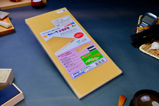 long yellow rectangular shaped rubber cutting board with product paper in hiragana showing bright green red blue images surrounded by kitchenware on grey background
