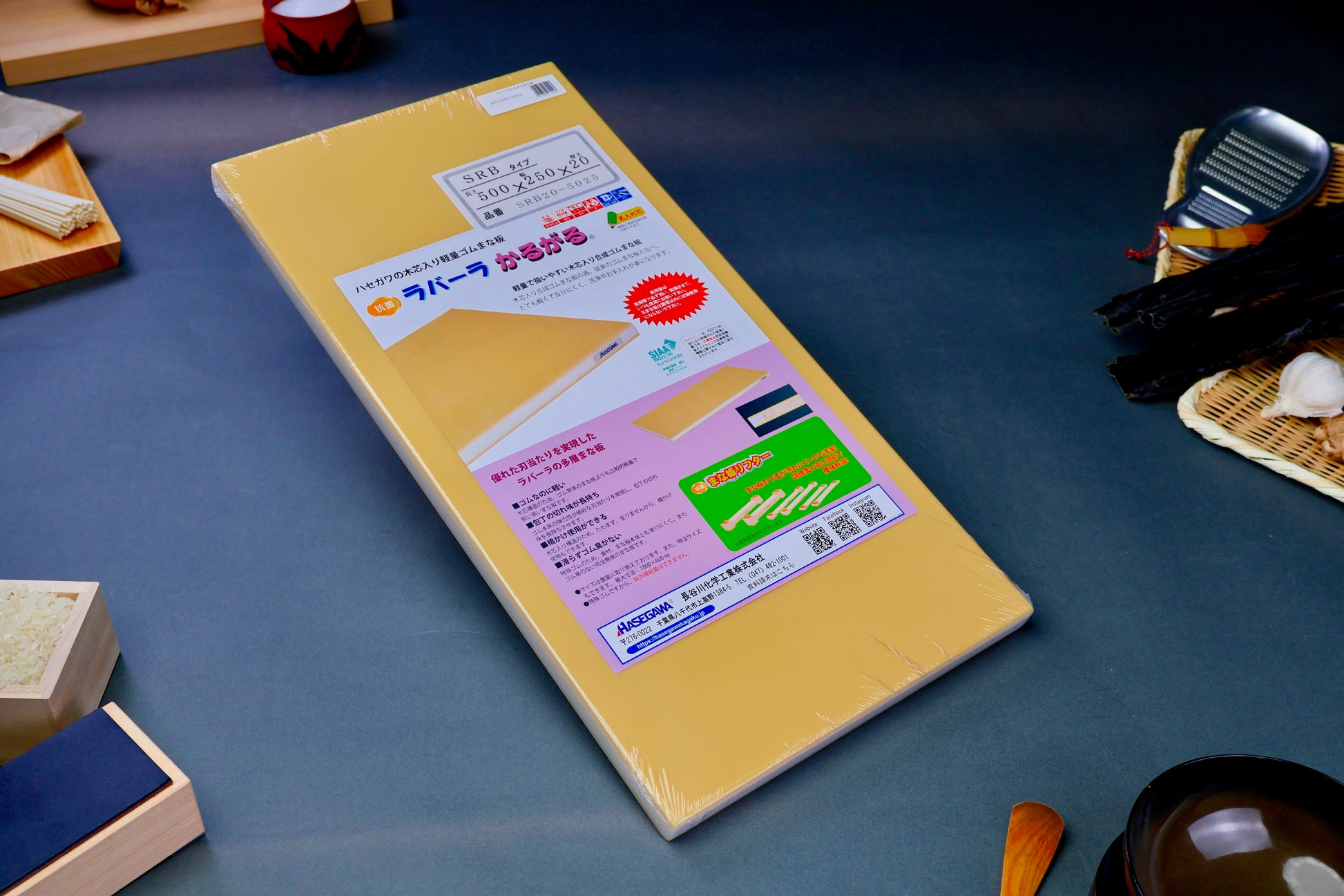 long yellow rectangular shaped rubber cutting board with product paper in hiragana showing bright green red blue images surrounded by kitchenware on grey background