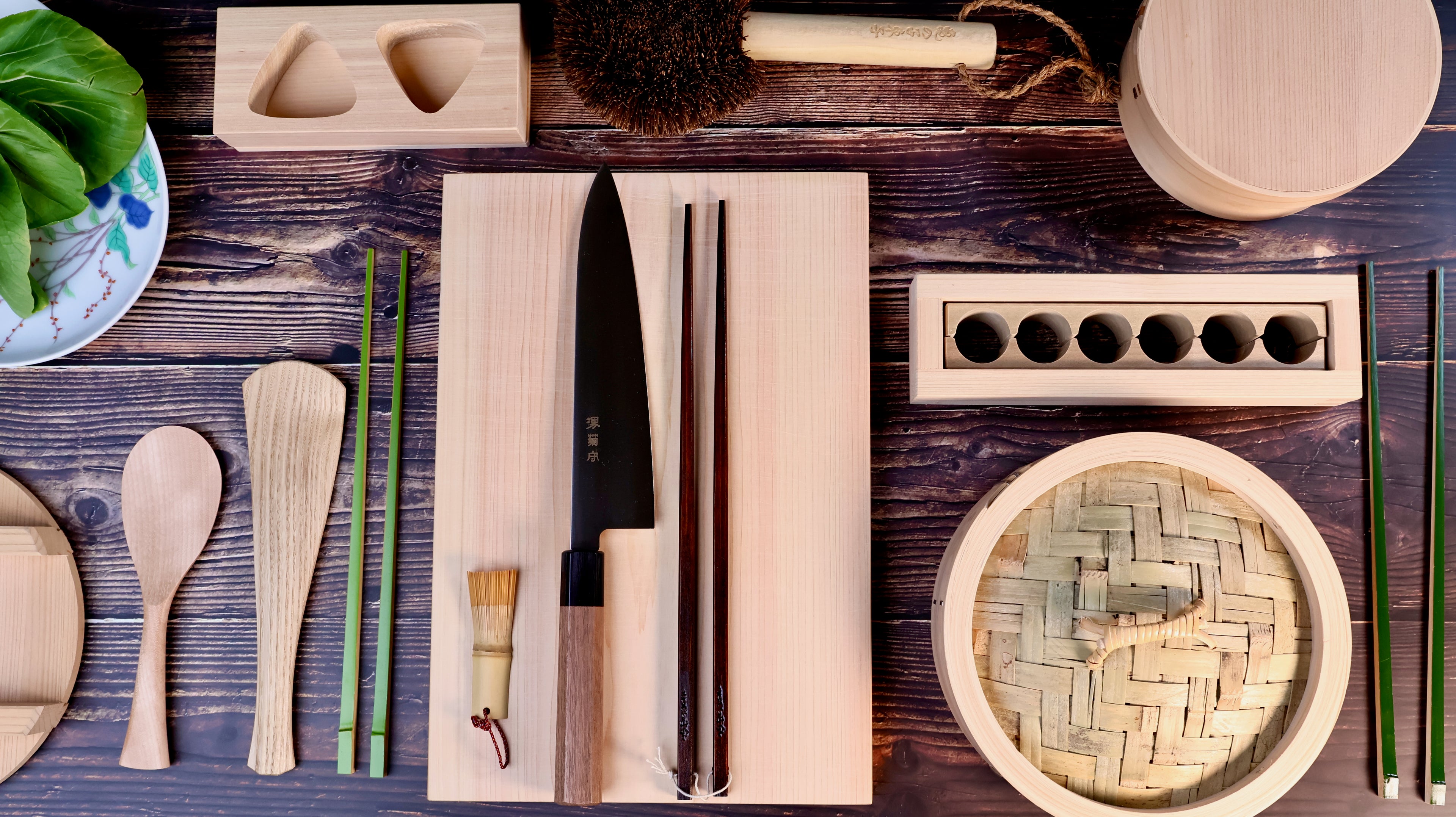 Overhead flat lay of Japanese kitchen tools on dark wood including black gyuto on hinoki board, bamboo steamer, wooden rice paddle and chasen brush