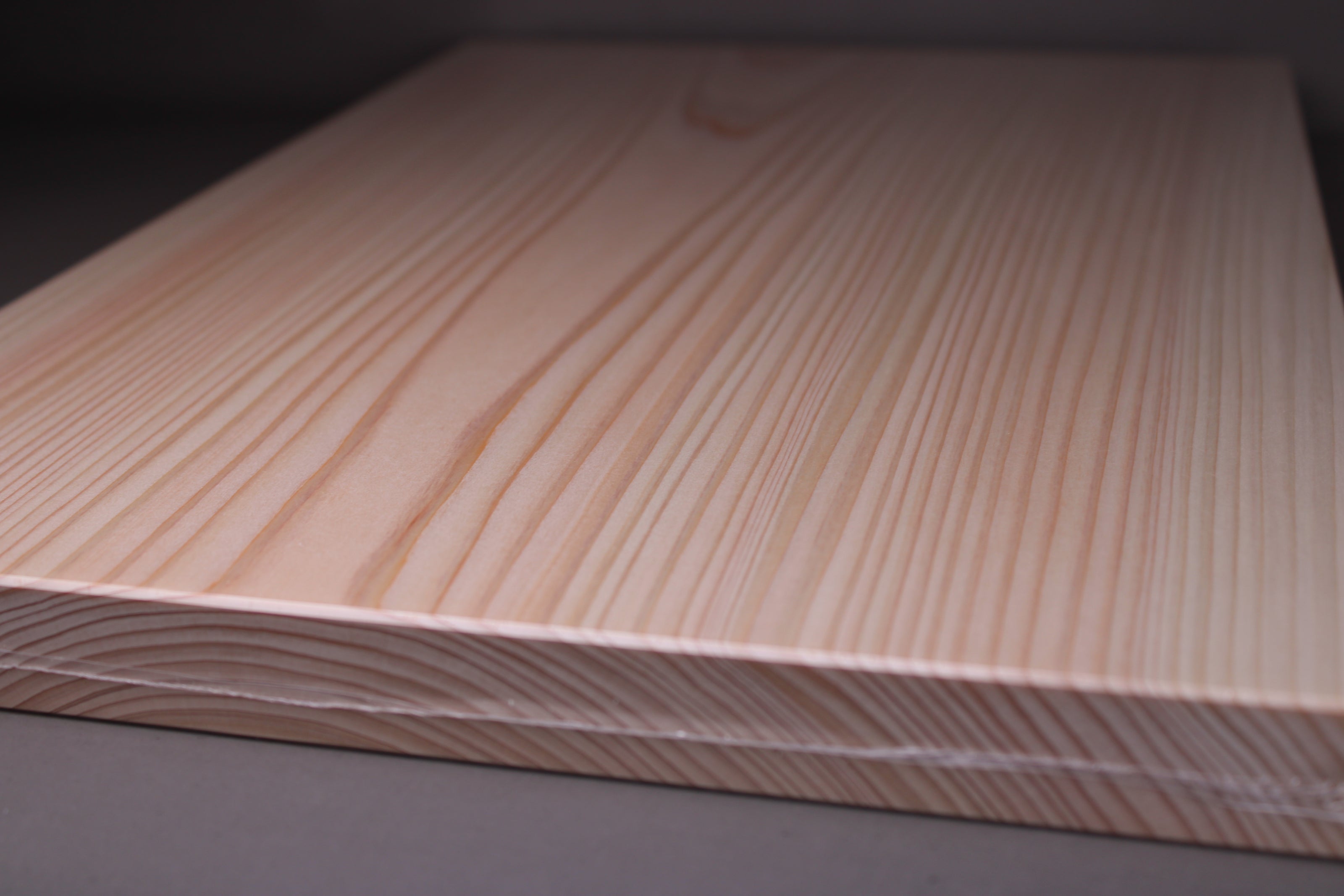 close up side of rectangular shaped light medium deep wood tan hinoki cutting board with deep natural grain lines and rings in front of dark grey backdrop