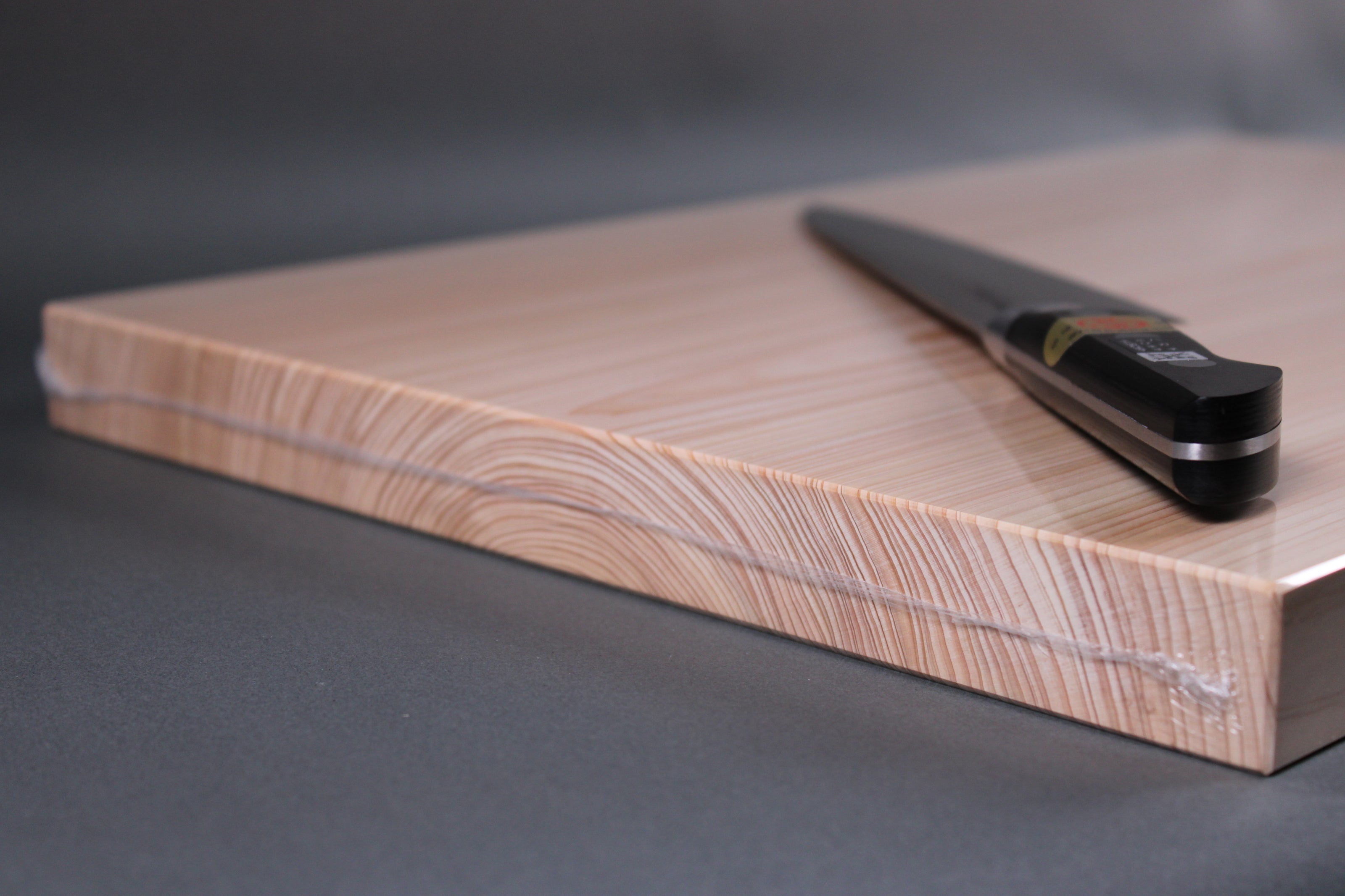 side detail of rectangular shaped light medium deep wood tan hinoki cutting board with deep natural grain rings with knife atop board in front of dark grey backdrop