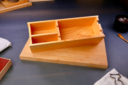 hinoki wood rectangular shaped box with natural wood grain sits atop wood cutting board surrounding japanese kitchenware on gray background