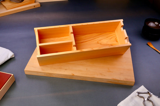 hinoki wood rectangular shaped box with natural wood grain sits atop wood cutting board surrounding japanese kitchenware on gray background