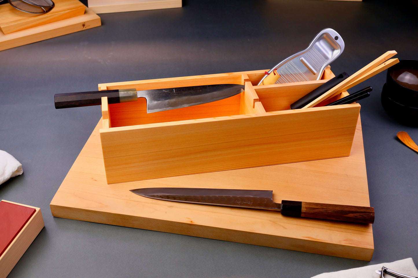 hinoki wood rectangular shaped box with natural wood grain containing knives and tools sits atop wood cutting board surrounding japanese kitchenware on gray background