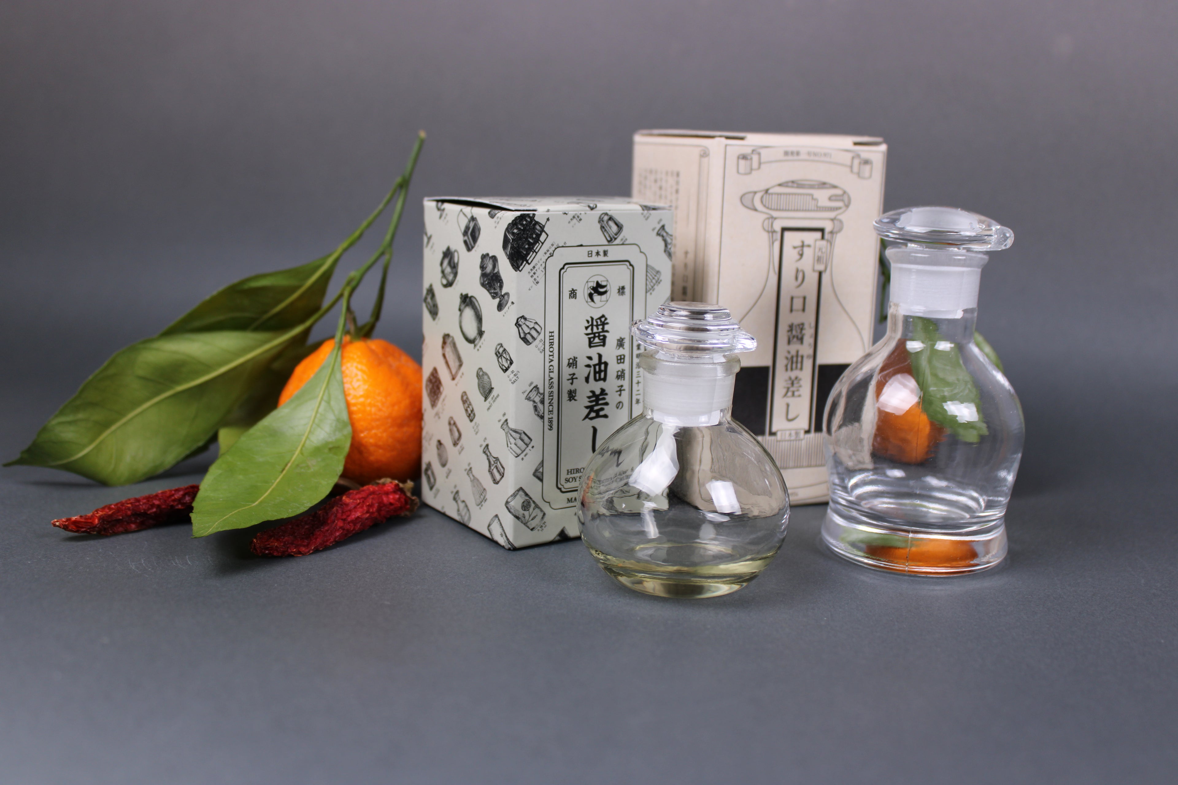 small and large Hirota glass soy sauce cruets in front two product boxes fruit dried pepper grey background