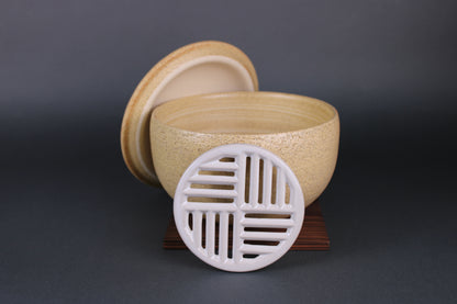 product image of clay earthenware vessel shaped round with a coarse yellowish look comes with a lid of the same look and feel and a ceramic grate inside for separating water and food the grey background contrasts with the yellowish hue very well