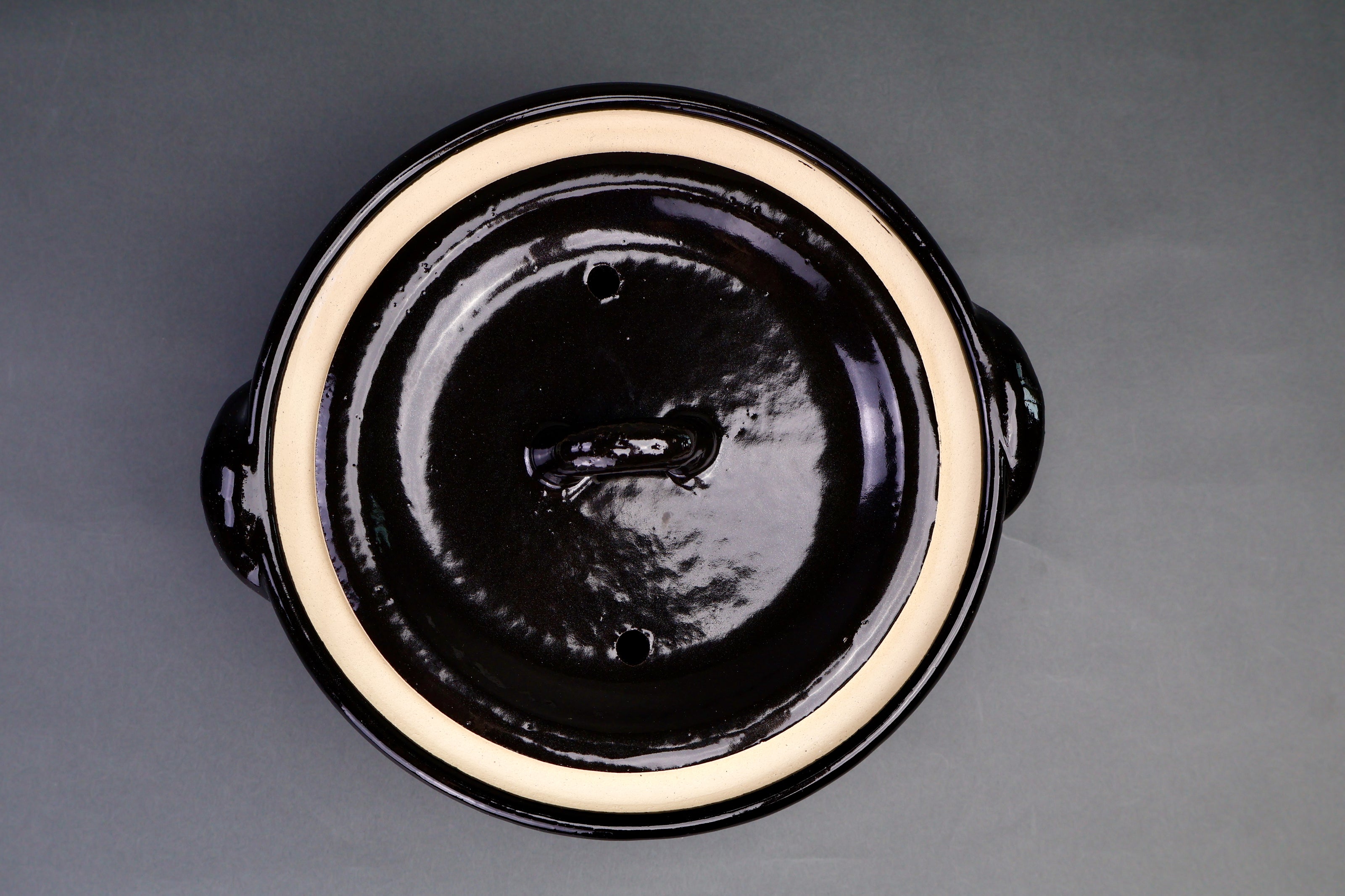 spotted black glazed rice donabe with shallow handles and slightly fluted lid surrounded by kitchenware and grey backdrop