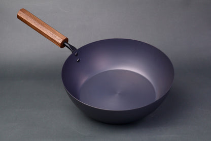 wide deep bluish black carbon steel pan long octagonal wood handle in light grey backdrop