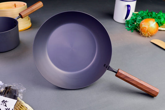 frontal shot of grayish black nitrided iron saute pan with brown wooden octagonal handle surrounded by Japanese kitchenware with light grey background